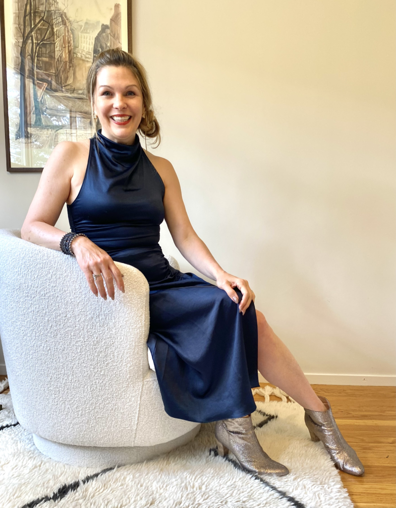 woman, Dr. Amy Backos smiling at the camera, wearing a blue dress and sitting in a white chair
